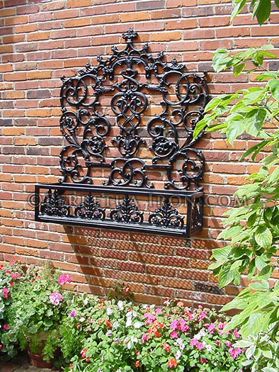 A Large scale wal mounted flowerbox mounted to a brick wall in a garden area.  Green plants surround the paislied and crolled cast iren components of this black iron and steel contruction.