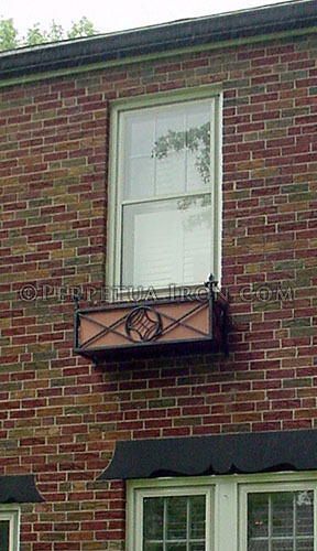 Fabricated steel flowerbox on a second story window viewed from ground level. This is a front view showing a single circle design framed by interconnecting X shapes.  The circle has a slightly concave, crossed diamond form suspended in the center. The background is a copper red panel and the corners are punctuated with fleur delis spikes rising above the box.