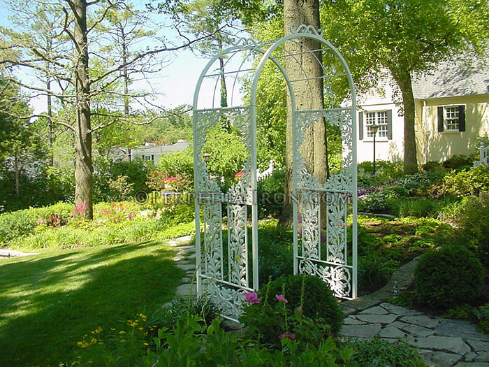 The is a white ornate decorative iron garden trellis archway with side panels and an open canopy over a flag stone walkway.