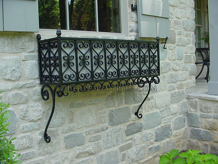 A high density scrolling designed black steel window box mounted below a window on a stone house. There are traditional painted gray shutters and muntined windows.