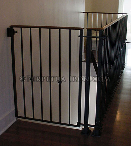 View of a custom decorative iron baby gate made to fit the top of stairs. The design is integrated into the existing handrail that wraps around an open stairwell hallway.
