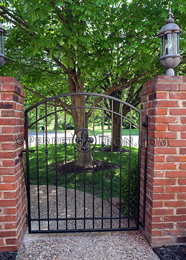 Custom iron arched gate with fleur de lis motif between two brick columns with loveyl trees in the background.