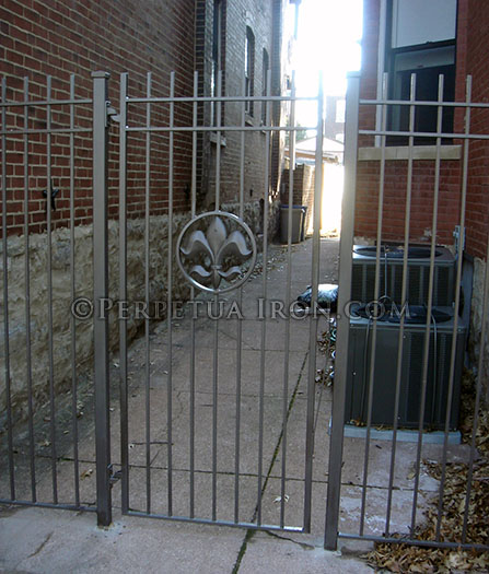 simple iron gate and panels with fleur de lis medallion between two old stone and brick houses.