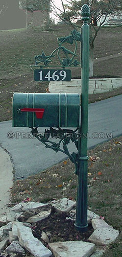 Mailbox post at the edge of a driveway surrounded by lanscaping stones.  The mailbox is green like aged copper and has organic leafy brackets where the house number hangs from above.